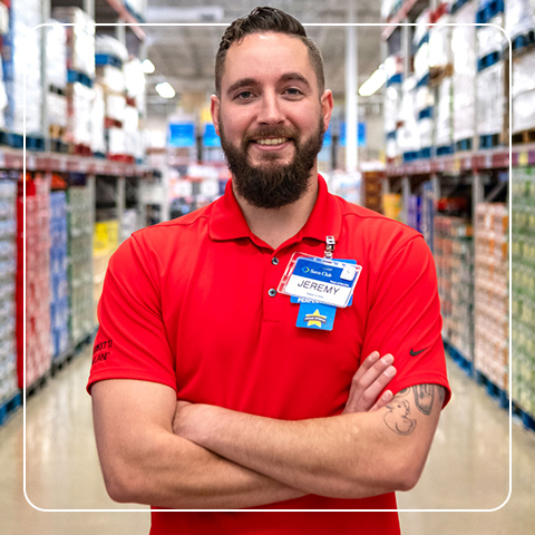 Smiling Sam's Club associate stands in a warehouse aisle, wearing a red vest and name tag.