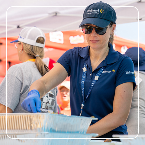 Walmart associate in blue vest and sunglasses serving food from a container at an outdoor event.