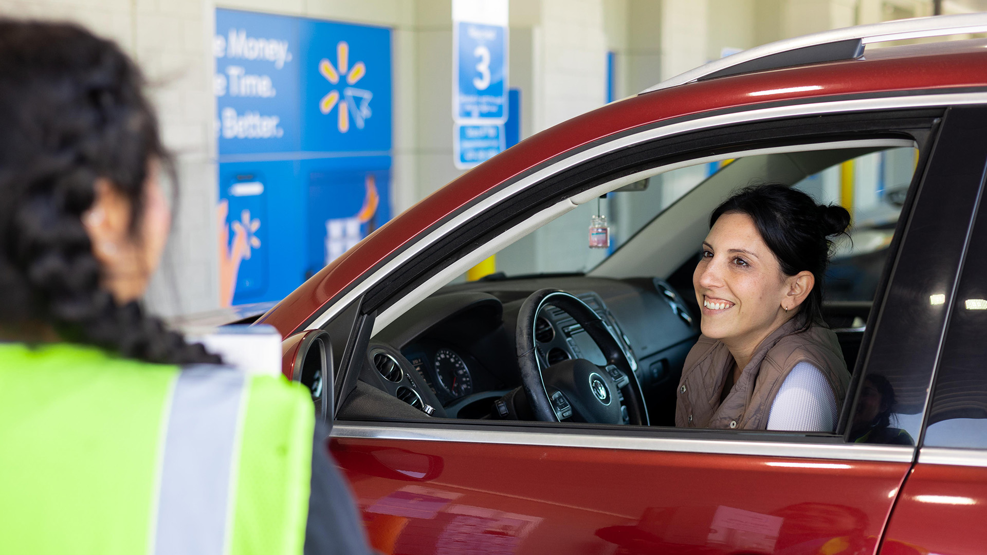 Walmart associate wearing a safety vest is helping a woman in a red car with their grocery pickup order.