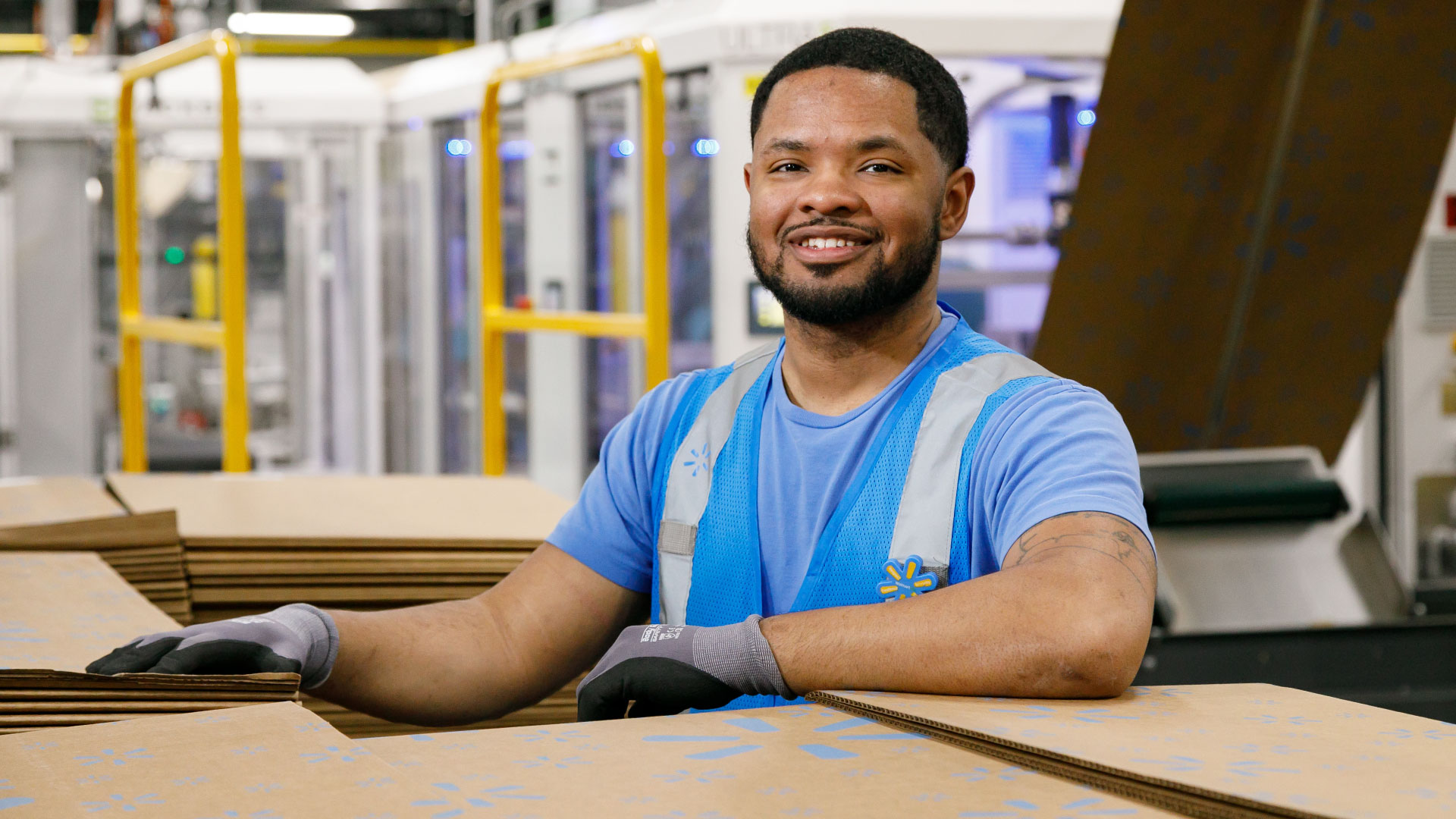 Smiling Walmart associate wearing a blue vest stands in a warehouse next to a stack of cardboard boxes.
