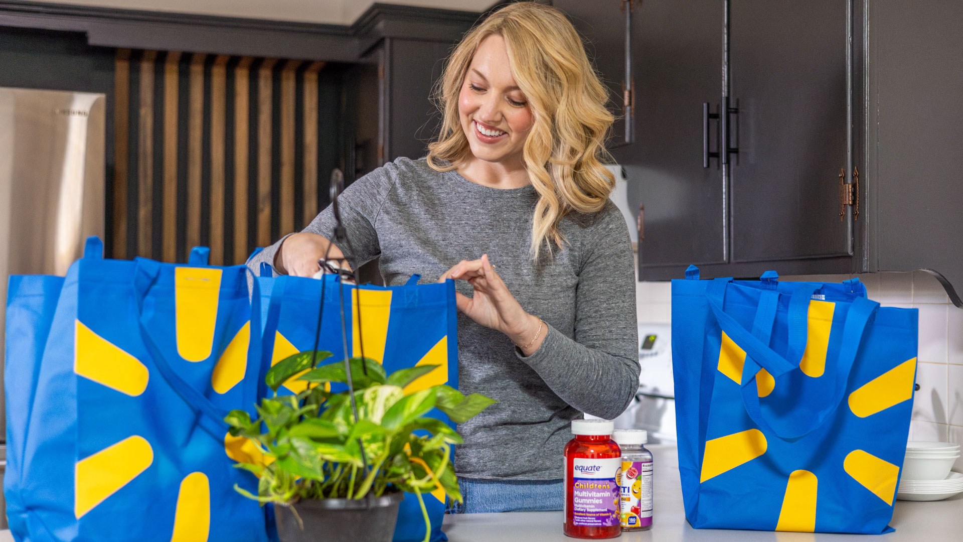 A woman wearing a gray shirt smiles while unpacking groceries from blue Walmart bags onto a kitchen counter.