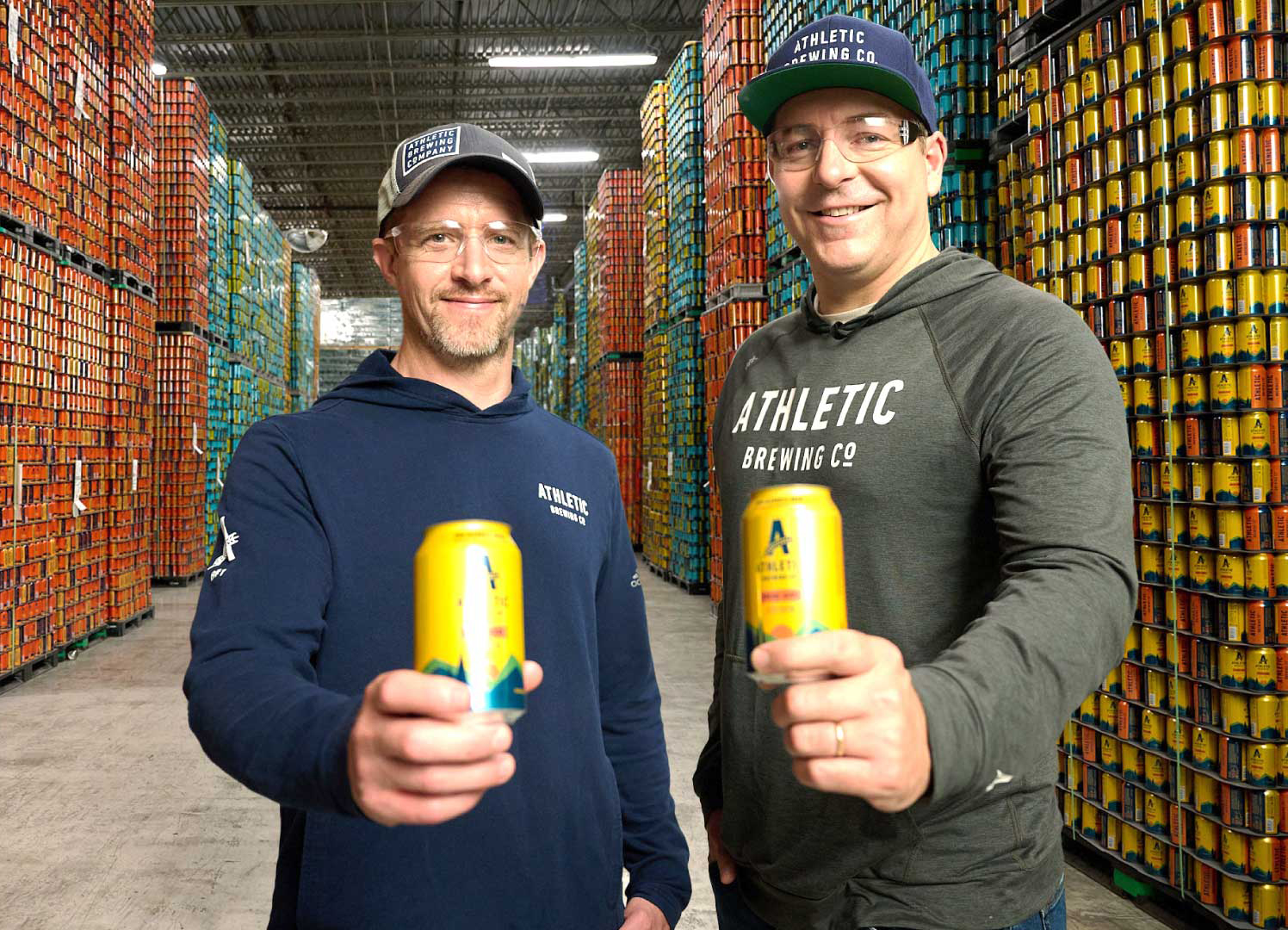 Owners of Athletic Brewing Co. hold up two cans of beer inside a warehouse.