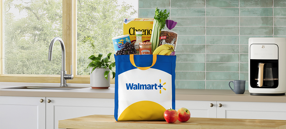A reusable Walmart+ grocery bag filled with various food items, including Cheerios cereal, bread, and fresh produce, placed on a kitchen counter. The kitchen features a modern design with a tiled backsplash, a sink, and a coffee maker.
