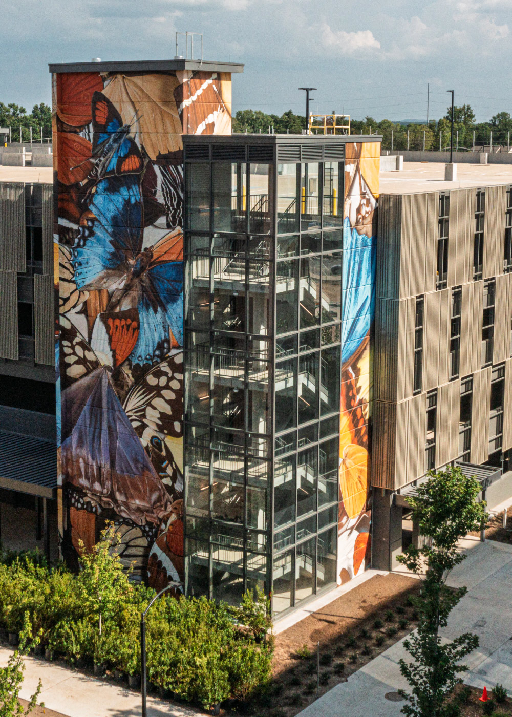 A vibrant mural featuring large, colorful butterflies adorns the side of a modern building with glass stairwell. The setting includes landscaped greenery and a clear sky, creating a lively urban environment. The artwork adds a striking visual element to the contemporary architecture.