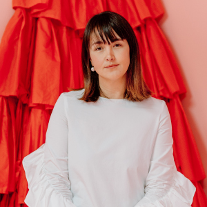 A woman wearing a white blouse with ruffled sleeves is positioned in front of a vibrant red backdrop featuring layered fabric. The setting emphasizes bold contrasts between the neutral clothing and the striking background. The image conveys a modern and stylish aesthetic with a focus on texture and color.