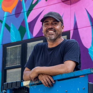 A vibrant mural featuring large, colorful flowers painted on a building wall. The artwork includes shades of purple, pink, orange, and green, creating a lively and artistic atmosphere. A person is standing on a blue platform in front of the mural, adding a sense of scale to the artwork. The setting appears to be outdoors during daylight.
