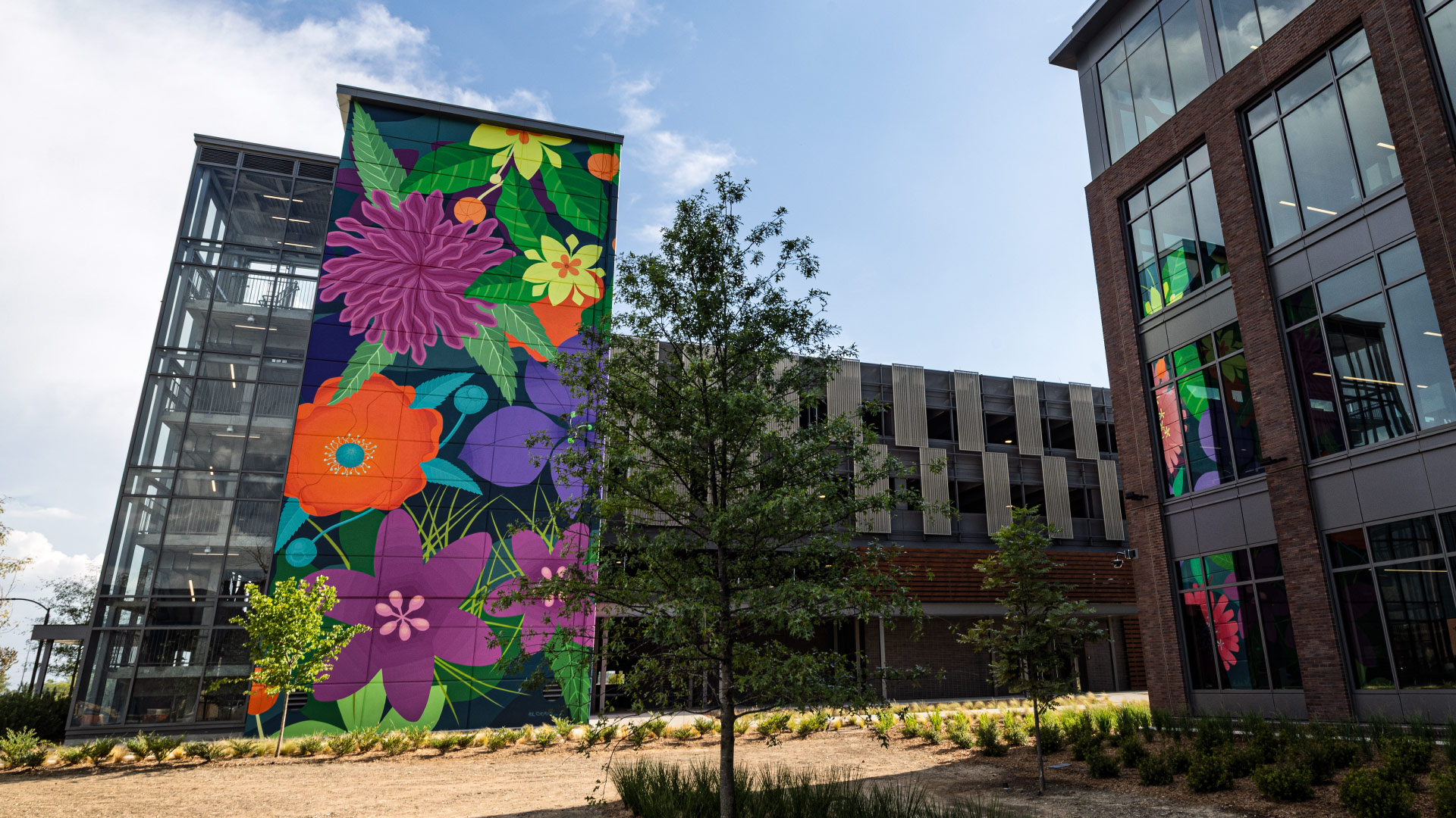 A vibrant mural featuring large, colorful flowers adorns the side of a modern glass and brick building. The setting includes landscaped greenery and a clear blue sky, creating a lively and inviting atmosphere. The artwork showcases bold hues of purple, orange, green, and yellow, adding a dynamic visual element to the urban environment.