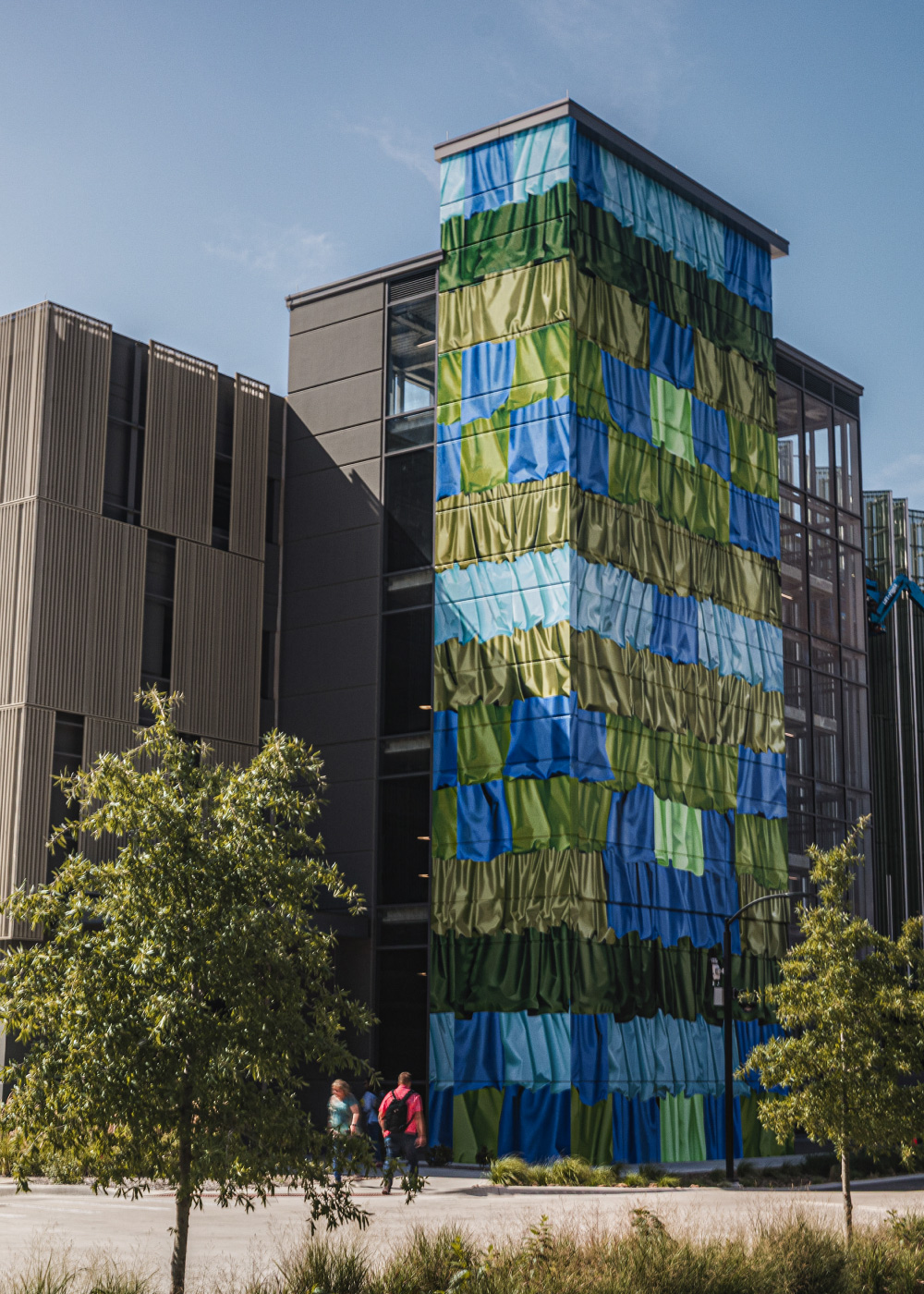 A striking vertical fabric art installation adorns the facade of a modern building. The patchwork design features vibrant shades of green, blue, and teal, creating a dynamic visual effect. The setting includes trees and pedestrians walking nearby, suggesting an urban environment. The artwork contrasts beautifully with the neutral tones of the building's architecture.