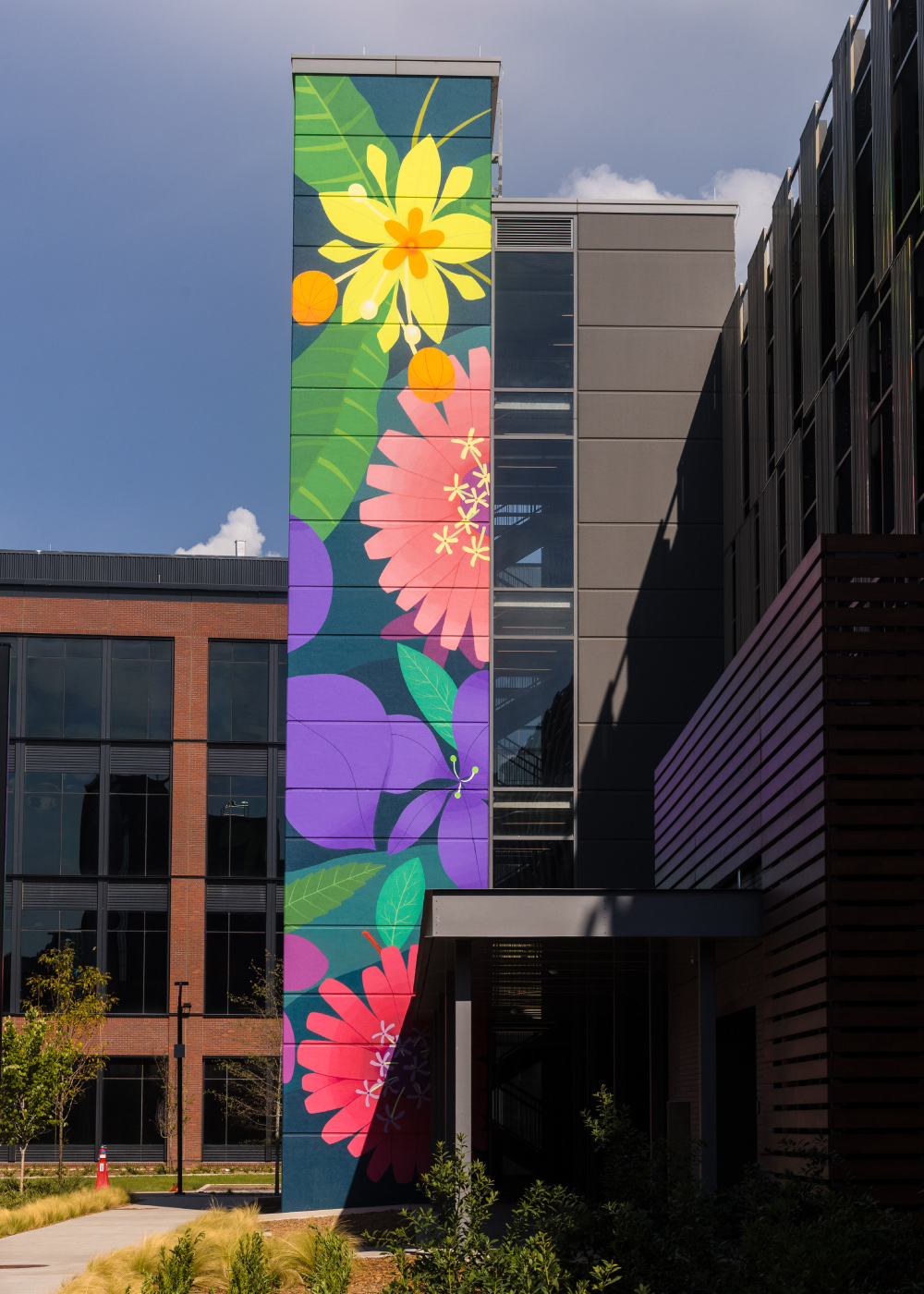 A vibrant mural featuring large floral designs in yellow, pink, purple, and green adorns the side of a modern building. The setting includes contemporary architecture with glass windows and wooden accents, surrounded by landscaped greenery. The artwork adds a lively and artistic touch to the urban environment under a partly cloudy sky.