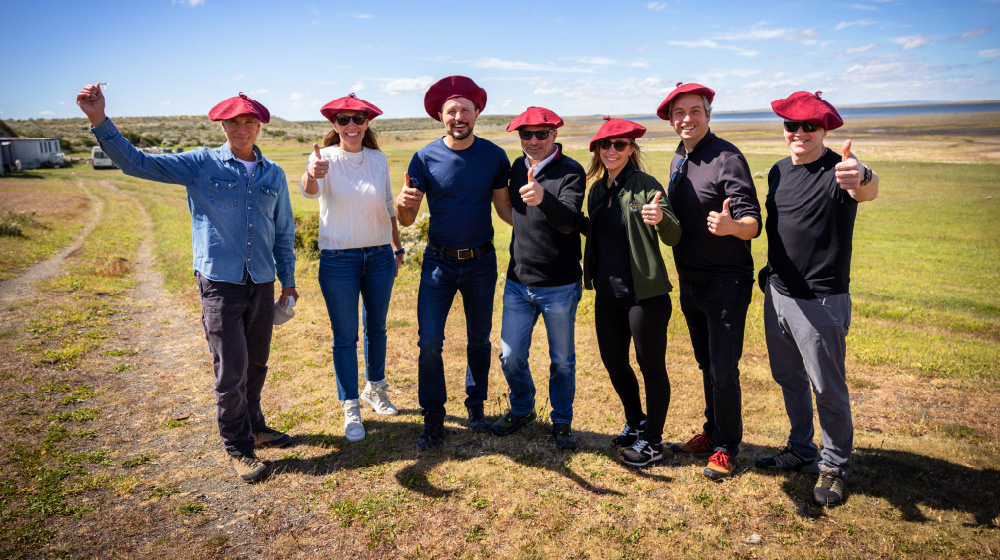 Kath McLay poses with six people in a grassy field. All are wearing red berets and giving a thumbs up.