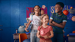 A group of children stands in a classroom setting with vibrant educational decor emphasizing a back to school theme. The background features a blue wall adorned with posters.