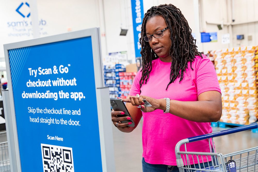 A woman scans a QR code on her phone at a Sam's Club. The sign promotes the Scan & Go checkout feature, allowing customers to skip the checkout line.
