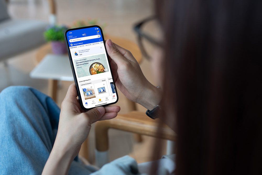 A person holds a smartphone displaying the Walmart mobile app interface while seated in a modern setting. The screen shows a food-related image and various app navigation elements. The individual is wearing casual clothing, and the background includes a blurred table and potted plant.