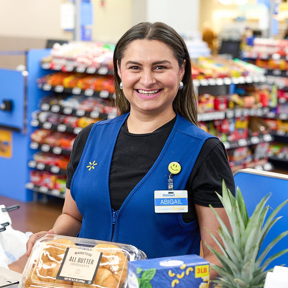 Smiling Walmart associate Abigail wears a blue vest and assists at the checkout counter. The setting includes a well-stocked store with visible shelves of products. Items on the counter include a pineapple, baked goods, and other groceries. The scene captures a typical shopping experience.