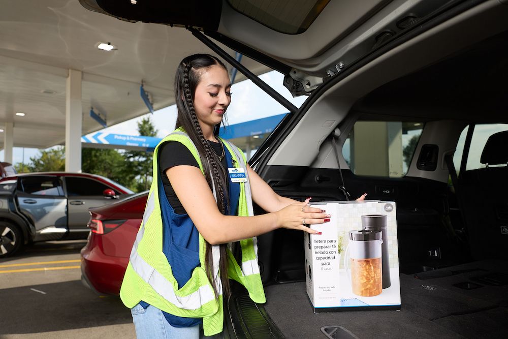 Walmart associate Brianna wears a high-visibility vest and loads a boxed kitchen appliance into the trunk of a vehicle. The box features an image of the product and visible text in Spanish. Cars and a canopy are visible in the background.