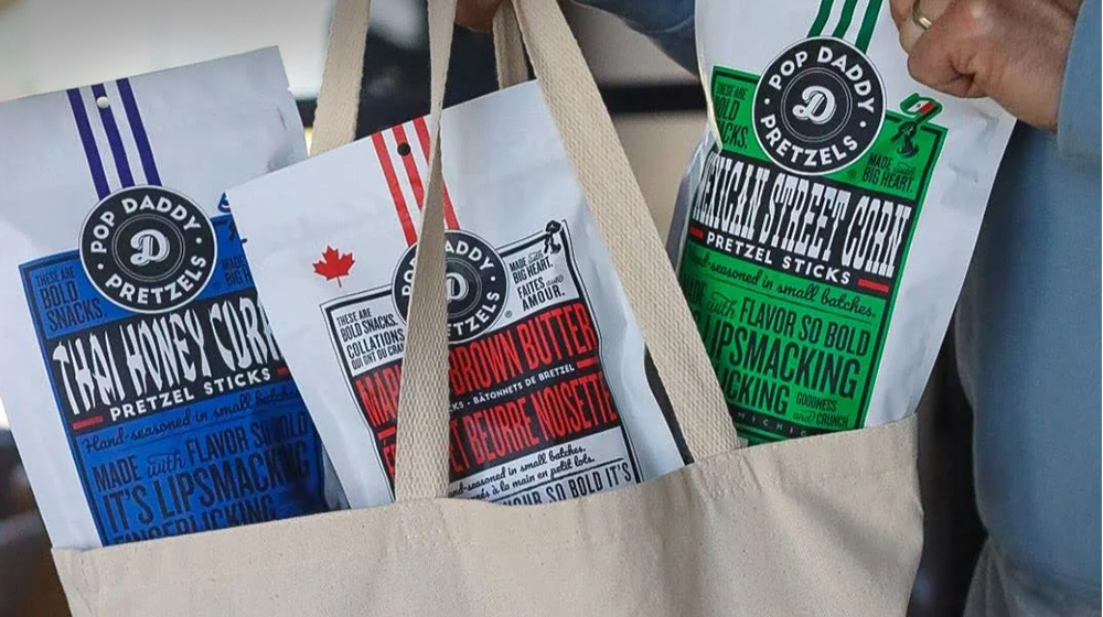 Three bags of Pop Daddy Pretzels are displayed in a beige tote bag. The flavors visible include 'Zesty Honey Corn,' 'Butter & Garlic,' and 'Yellow Cheddar Cheese Corn.' 