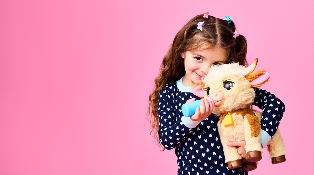 A young girl is holding a plush toy resembling a cow while standing against a vibrant pink background. She is wearing a navy blue outfit with white heart patterns and has colorful hair accessories.