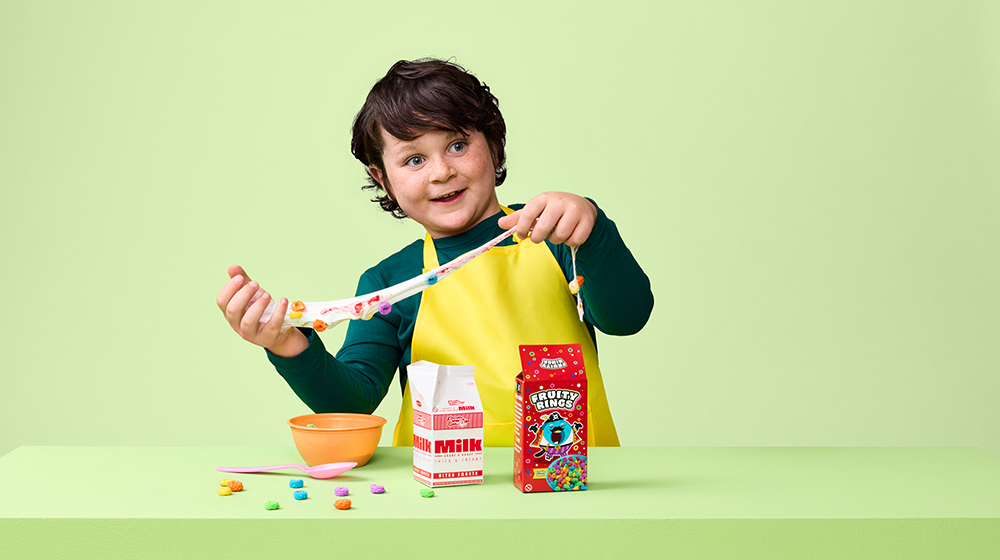 A child wearing a yellow apron is creating candy slime using colorful sweets on a light green background. The setup includes a red candy pack, a white and red ingredient bag, and a small orange bowl. 