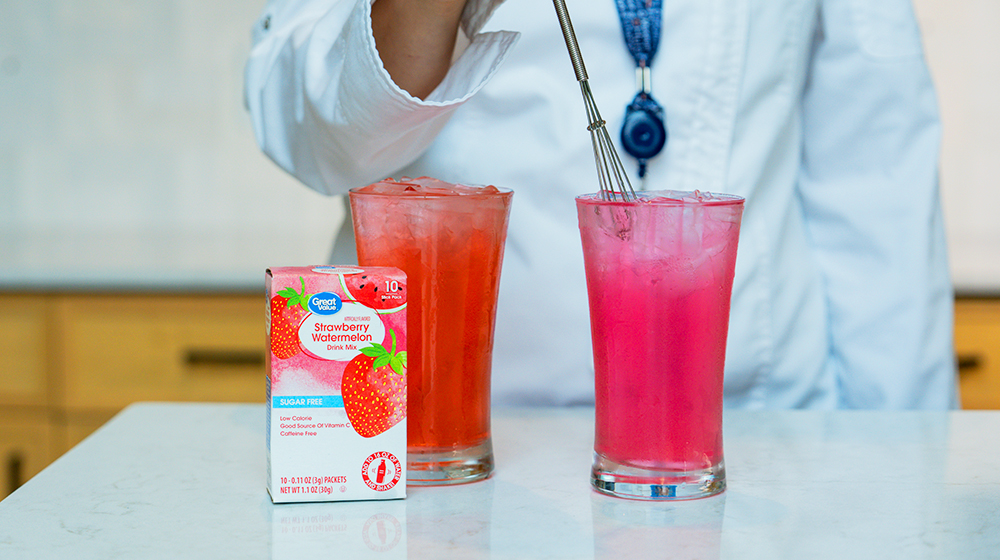 A box of strawberry drink mix is displayed alongside two vibrant iced beverages on a countertop. The drinks feature pink and red hues, with one being stirred using a metal whisk. The setting appears to be a kitchen, with a person partially visible wearing a white shirt and blue necklace.