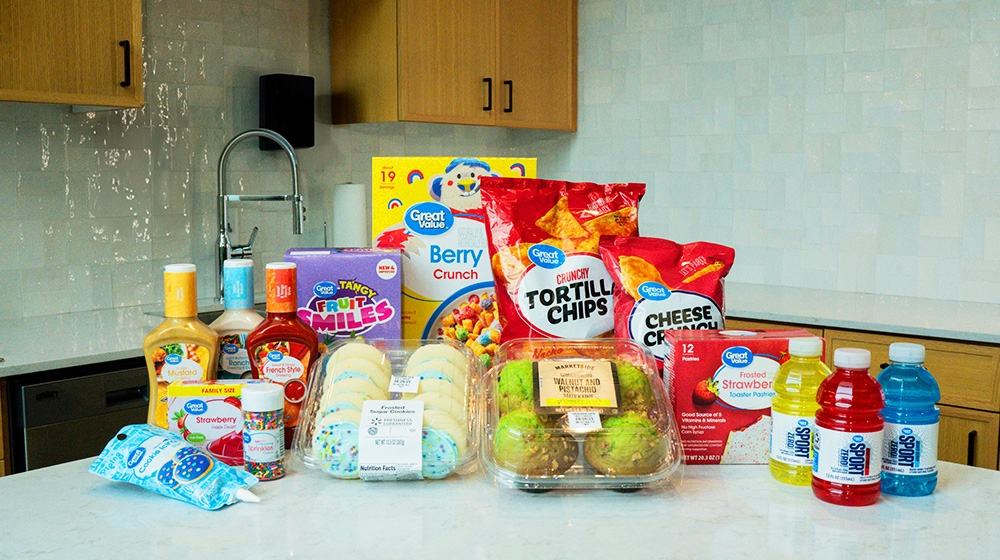 A variety of grocery products displayed on a white kitchen counter, including snacks, beverages, and condiments. Visible items include tortilla chips, cookies, sports drinks, and salad dressings. Packaging features clear branding and text such as 'Great Value' and 'Marketside.'