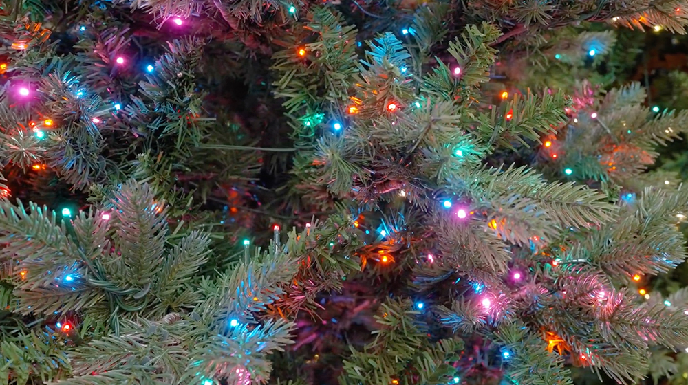 A close-up view of a holiday tree adorned with vibrant multicolored lights. The evergreen branches are illuminated with hues of blue, red, orange, and pink, creating a festive and cheerful atmosphere. The image captures the essence of holiday decor and celebration.