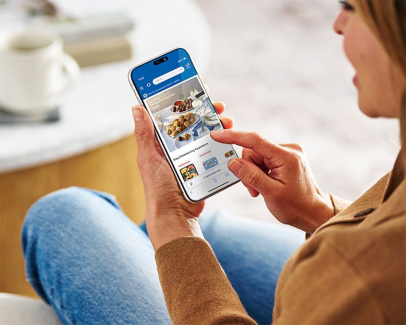 A person holds a smartphone while browsing the Sam’s Club app. The setting appears to be a cozy indoor space with a coffee table and books in the background. The individual is wearing casual clothing, including a brown sweater and blue jeans.