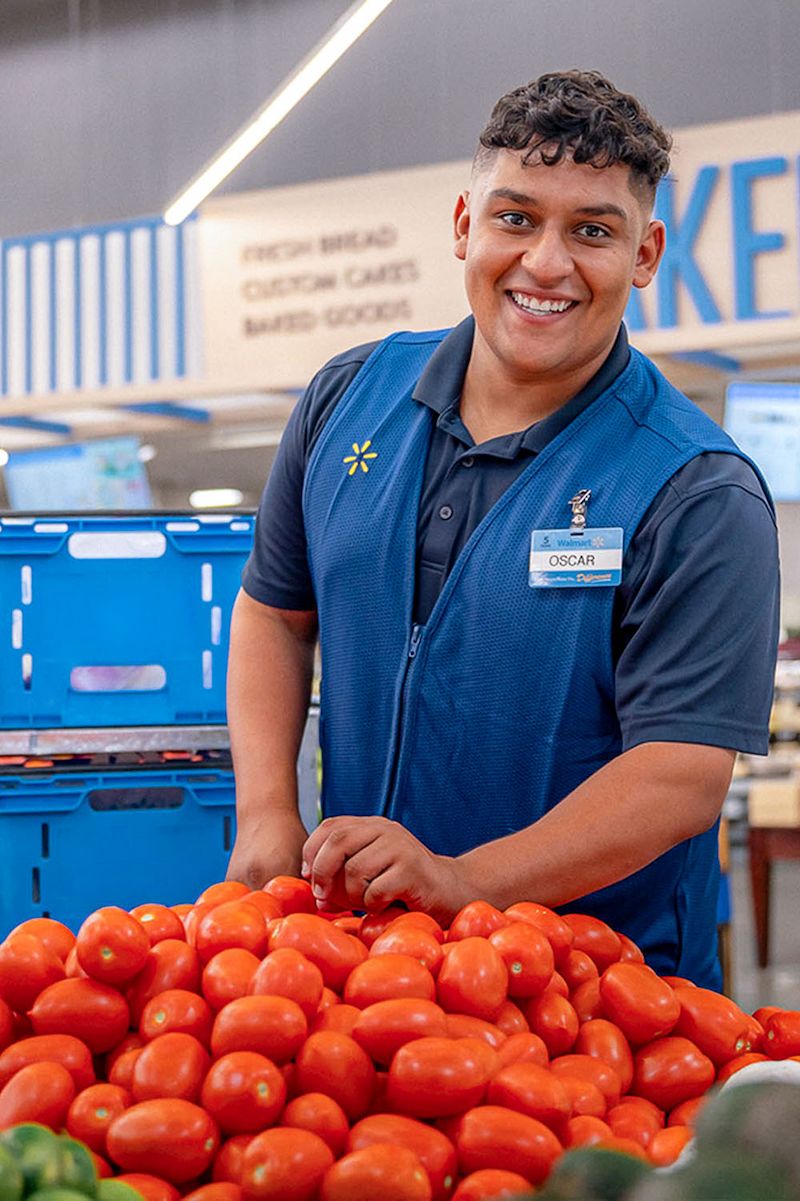 Walmart associate Oscar is wearing a navy blue shirt and a Walmart vest while selecting a tomato for a grocery order.