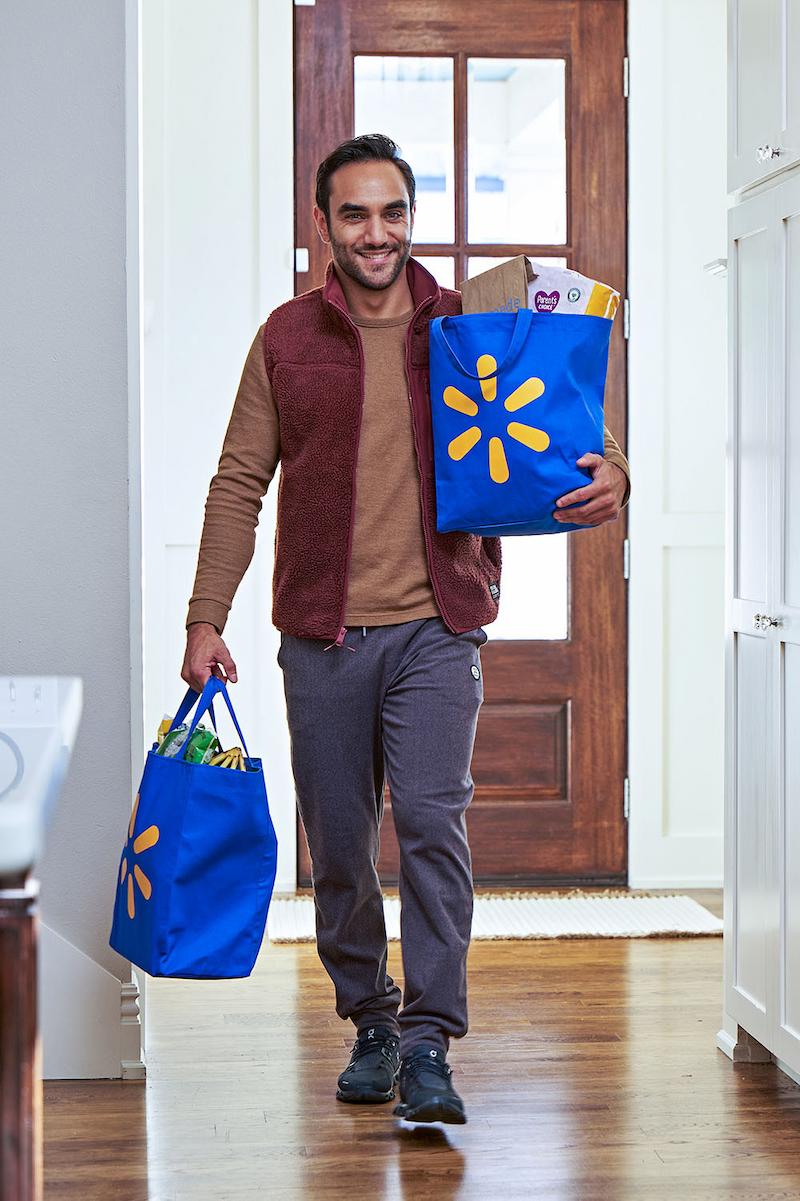 A Walmart customer is seen walking through a well-lit home interior carrying two full blue Walmart reusable shopping bags. The setting includes a wooden floor, white cabinetry, and a front door with glass panels.