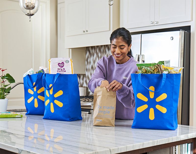 A Walmart customer in a purple sweatshirt is seen unpacking a prescription and groceries inside a kitchen. The kitchen features a modern design with white cabinets and a stainless steel refrigerator.