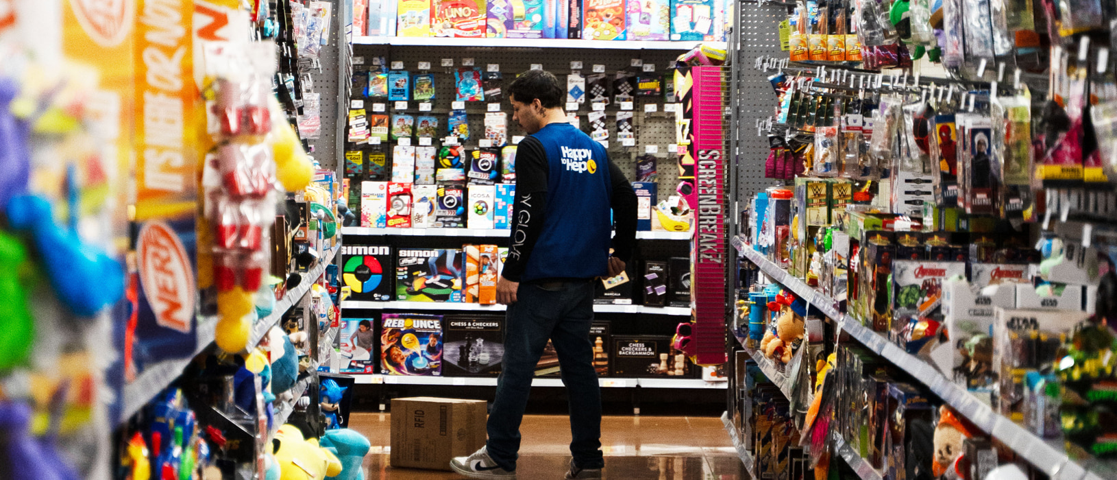 A Walmart associate wearing a blue vest is seen stocking the toy aisle. The shelves are filled with colorful toys, games, and action figures.