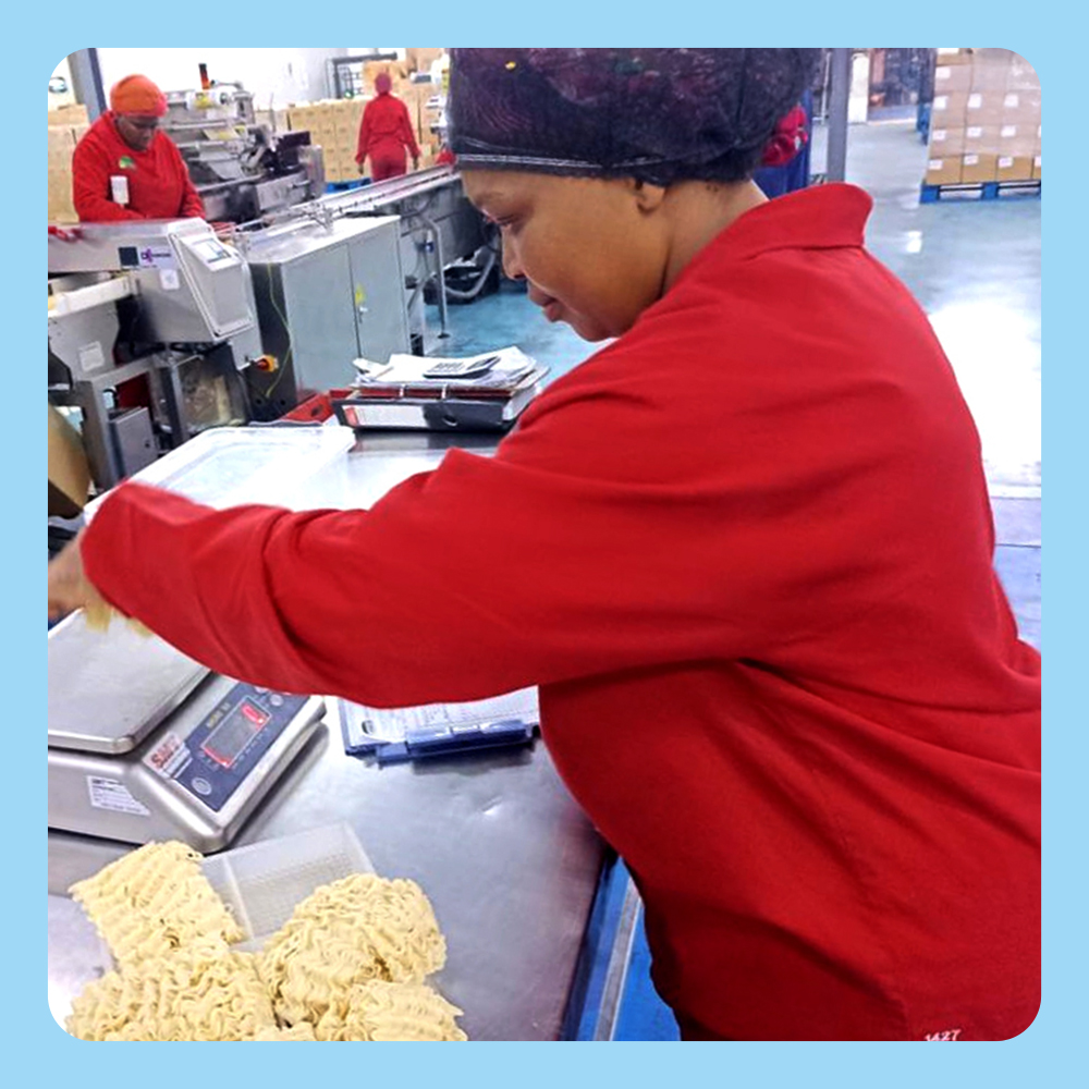 Nokuphumla Gqabi, wearing a red uniform, is seen weighing noodles on a digital scale in a factory environment. The setting includes industrial equipment and other workers in the background. The workspace is organized with visible packaging materials and machinery.