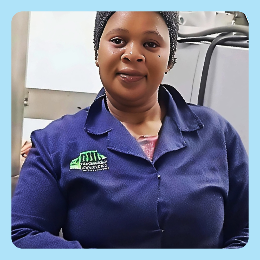 Nomahlubi Tshandana, wearing a blue uniform with an embroidered logo, stands near industrial machinery in The Noodle Factory. The uniform features a green and white embroidered design on the chest.