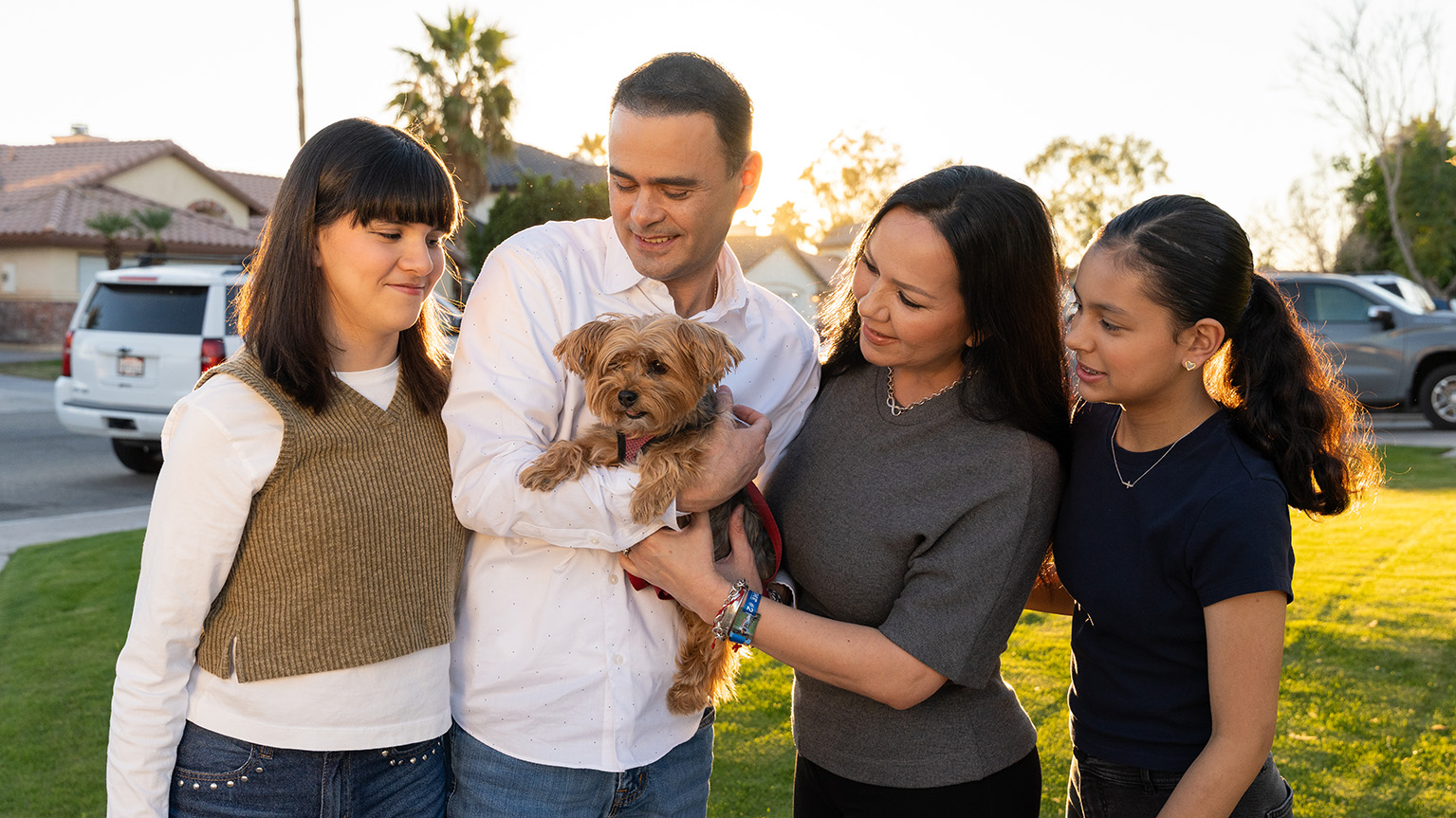 Oscar Romero and family stands closely together outside in a suburban neighborhood, holding a small brown dog.