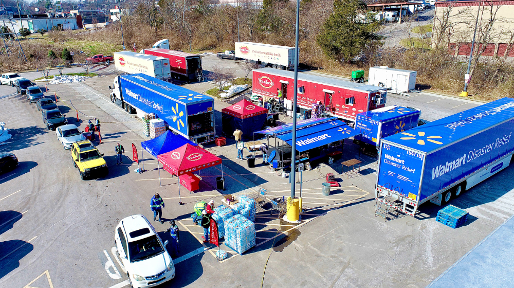 A fleet of Walmart Disaster Relief trucks is parked in an outdoor lot, distributing supplies to people in vehicles and on foot.