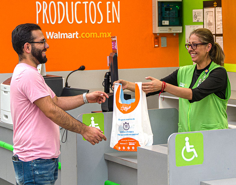 A customer at a Walmart checkout counter receives a shopping bag from an associate wearing a green vest.
