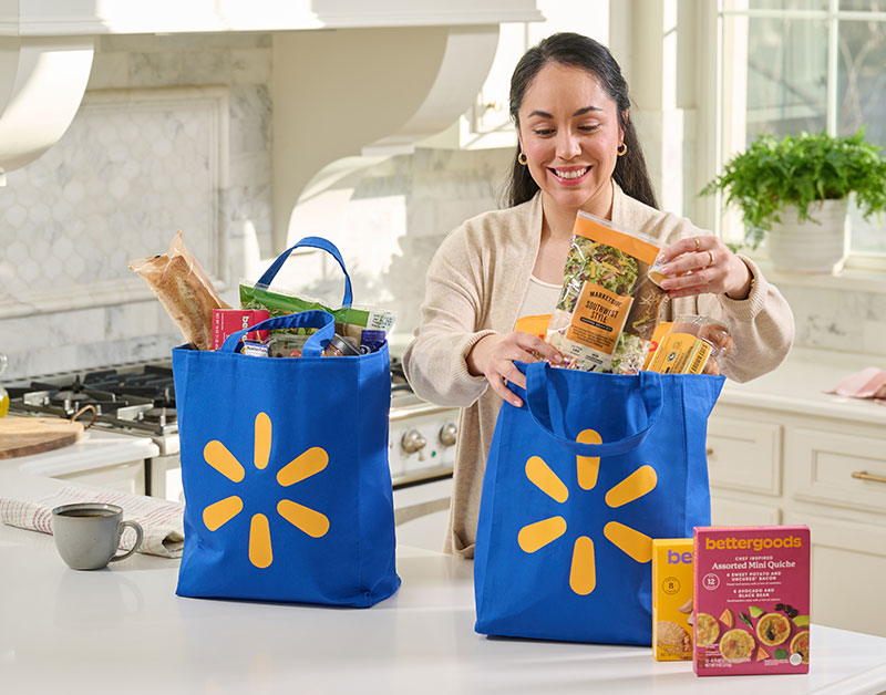 A woman stands in a bright, modern kitchen unpacking groceries from two Walmart reusable shopping bags.