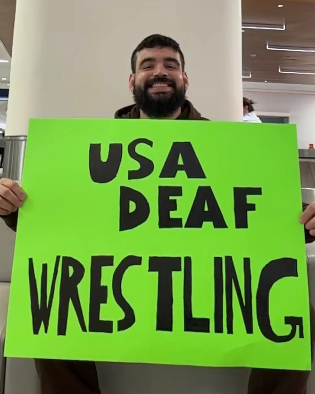 Harold Ramirez Jr. holding a bright neon green sign with bold black text reading 'USA DEAF WRESTLING.'