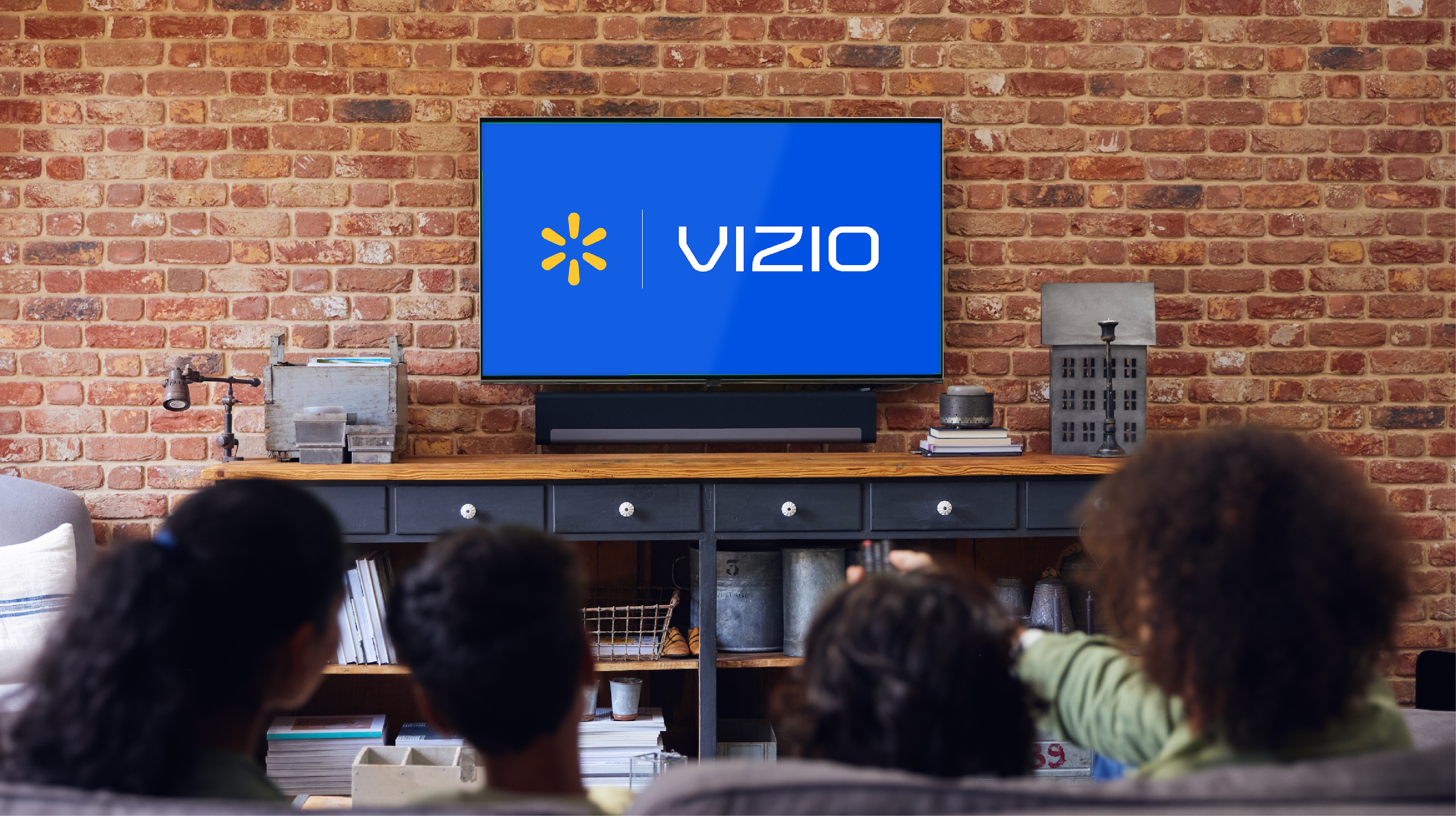 A group of four people sits together on a couch, watching a Vizio television in a home living room. The TV screen displays the unmistakable Vizio logo on a blue background. The setting features a brick wall, wooden console, and modern decor, creating a warm and inviting atmosphere. The group includes adults and children, viewed from behind, suggesting a family movie or TV night.