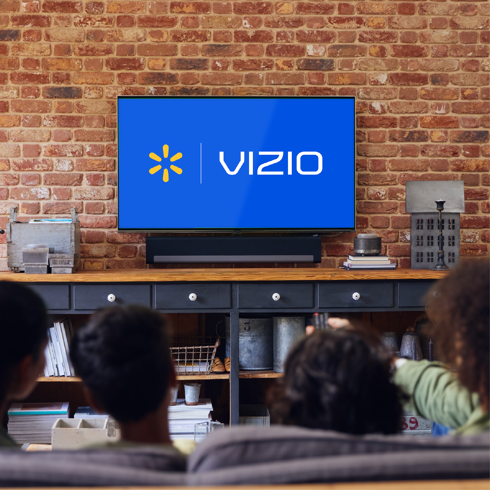 A group of four people sits together on a couch, watching a Vizio television in a home living room. The TV screen displays the unmistakable Vizio logo on a blue background. The setting features a brick wall, wooden console, and modern decor, creating a warm and inviting atmosphere. The group includes adults and children, viewed from behind, suggesting a family movie or TV night.