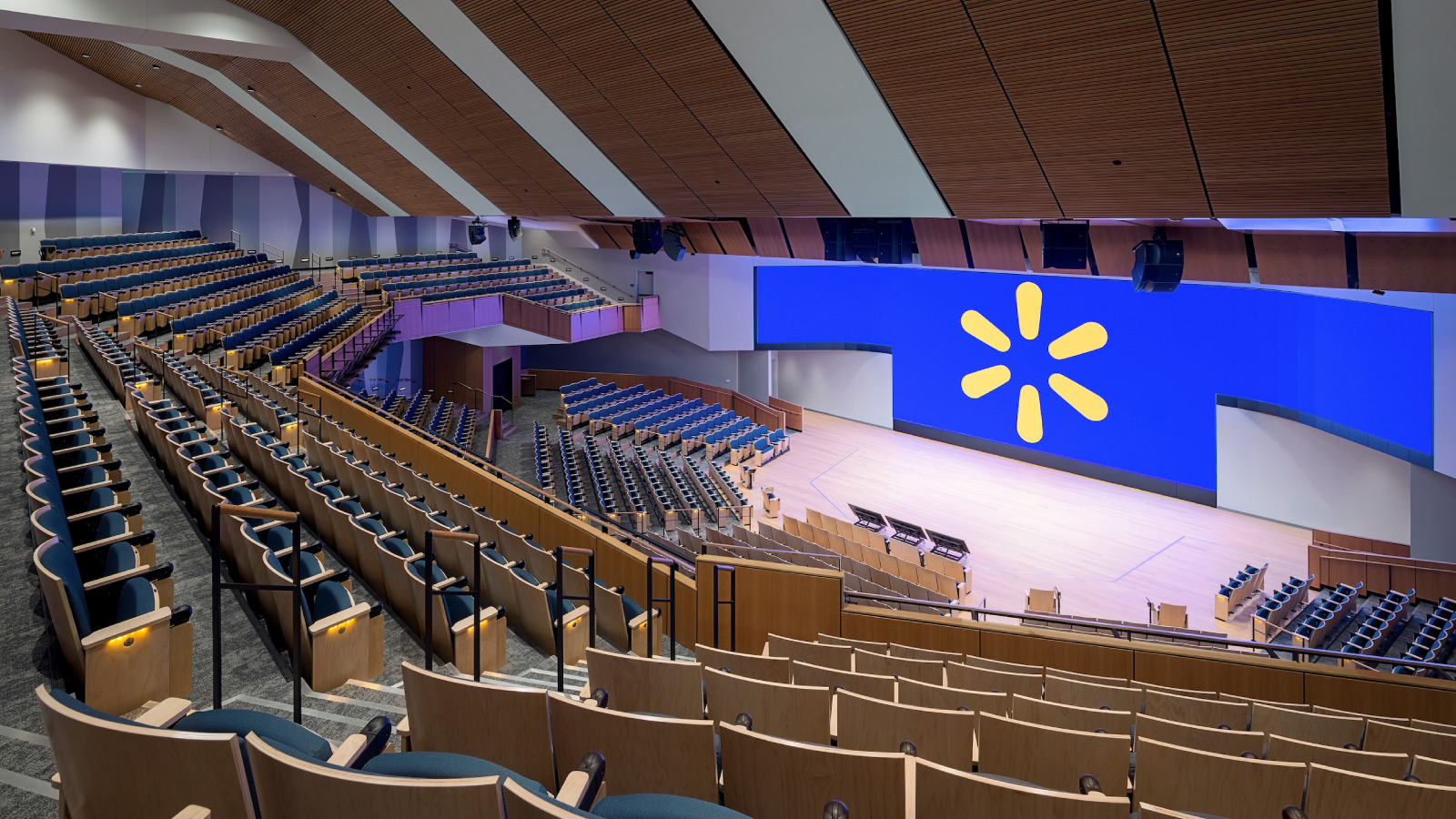 The auditorium in Sam Walton Hall, empty, with a large Walmart spark logo on the screen behind the stage.