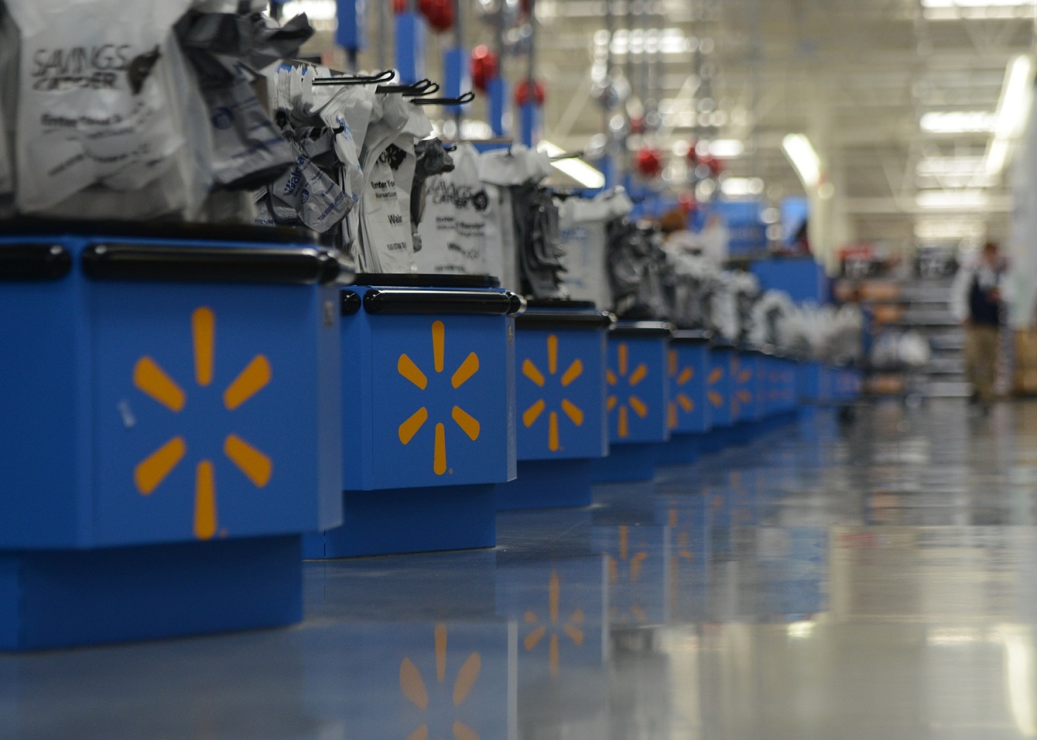 A long line of blue registers with Walmart's spark are stocked with empty plastic shopping bags. Two customers walk past the registers pushing their recently purchased items.