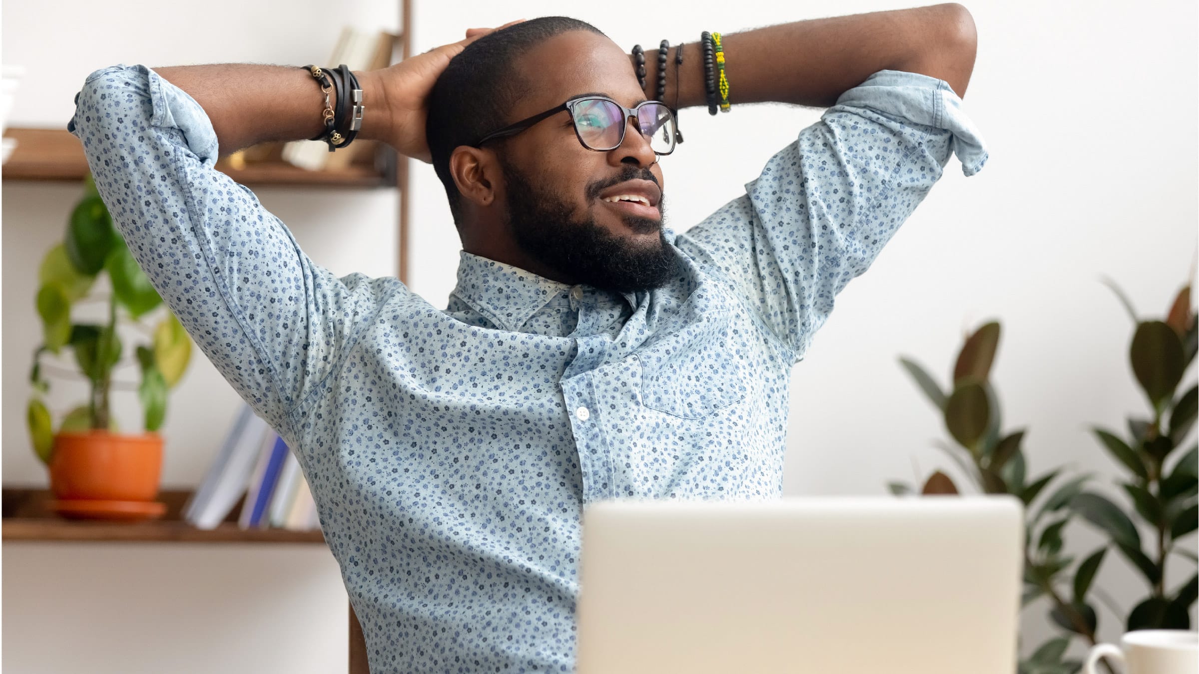 A man leans back in his chair away from his desk and laptop. His hands rest behind his head in a stretch.