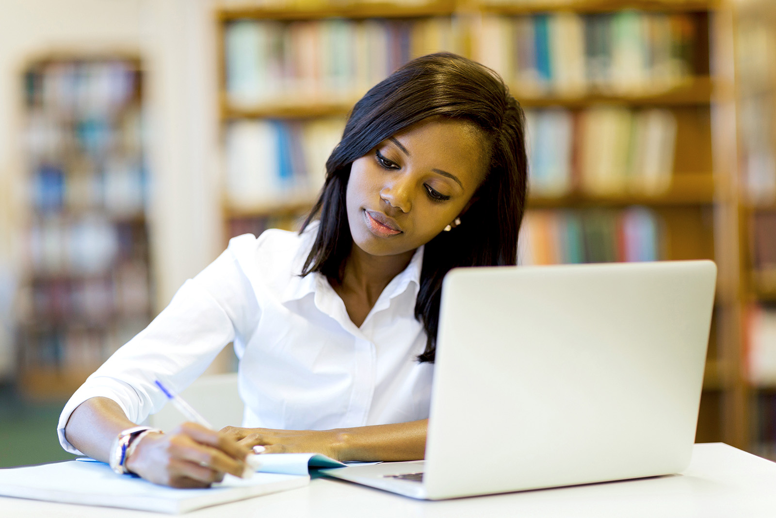 A woman sits at a desk with a notepad she's writing and an open laptop in front of her.