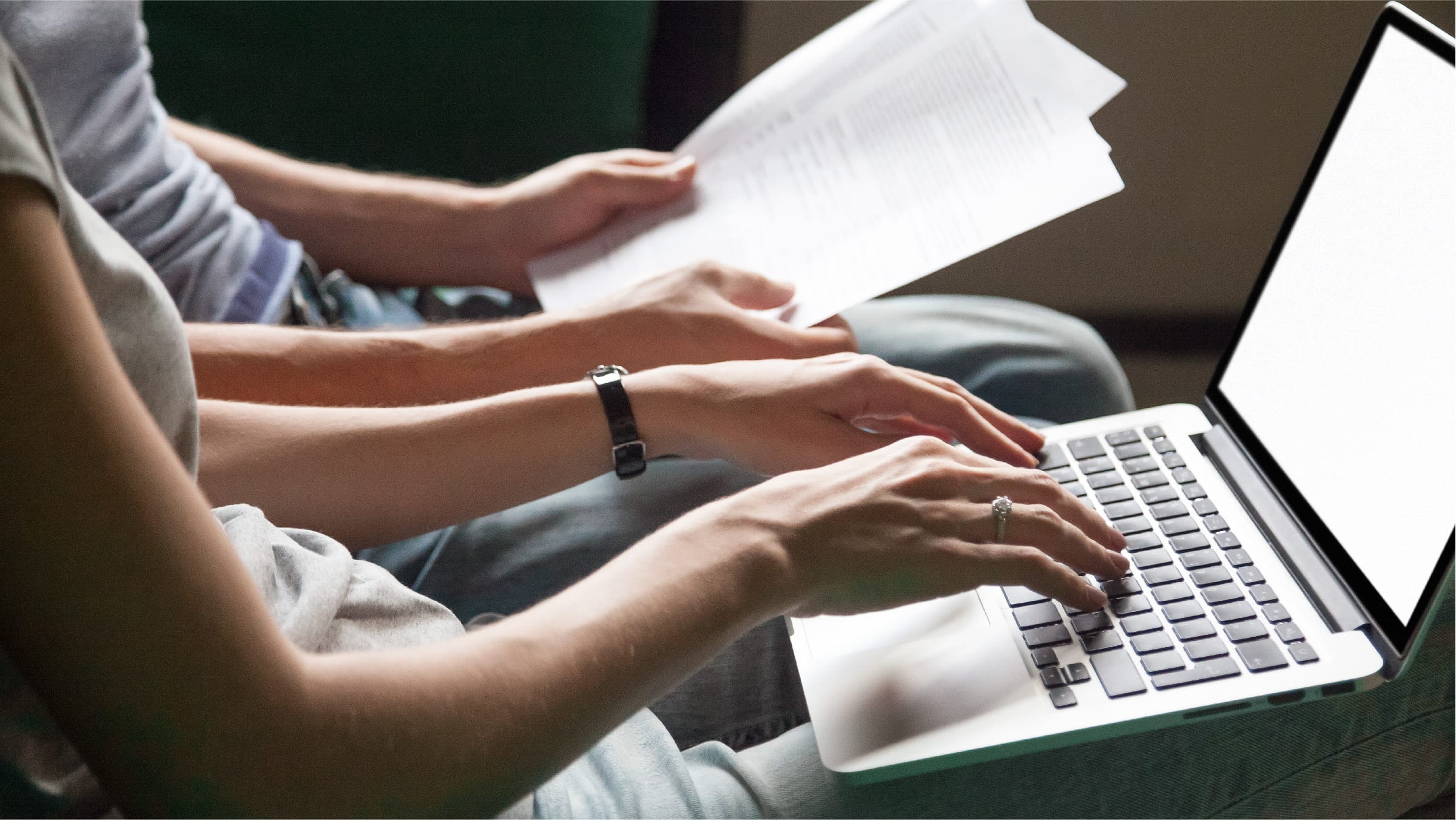 A woman works on a laptop in her lap while a man next to her looks through paperwork.