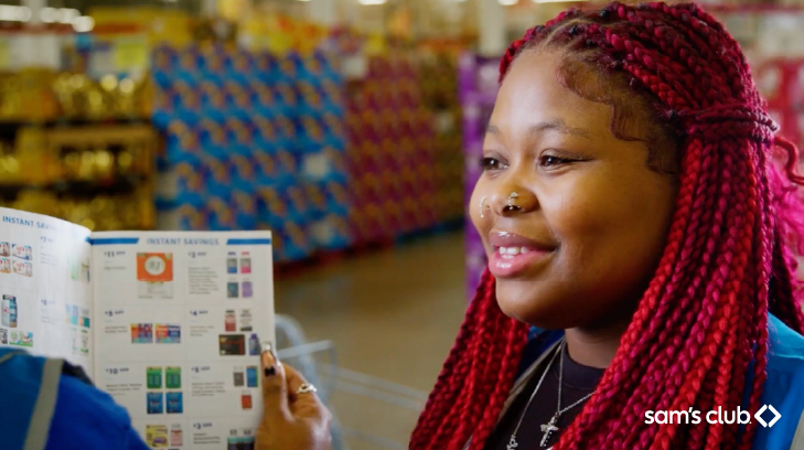A Sam's Club associate wearing a blue vest stands in the store smiling. The Sam's Club logo is in the bottom right corner.
